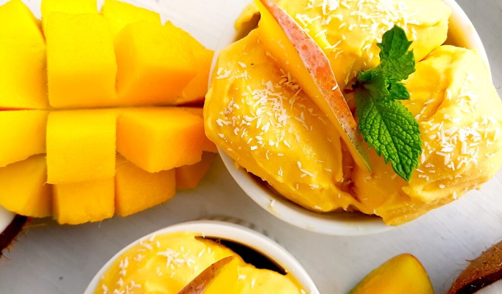 Homemade mango coconut soft serve ice cream in a white ramekin topped with shredded coconut and mint sprigs. Surrounded by halved coconut, sliced mangos and a second cup of ice cream. On a white background overhead shot. Sugar with Spice Blog.