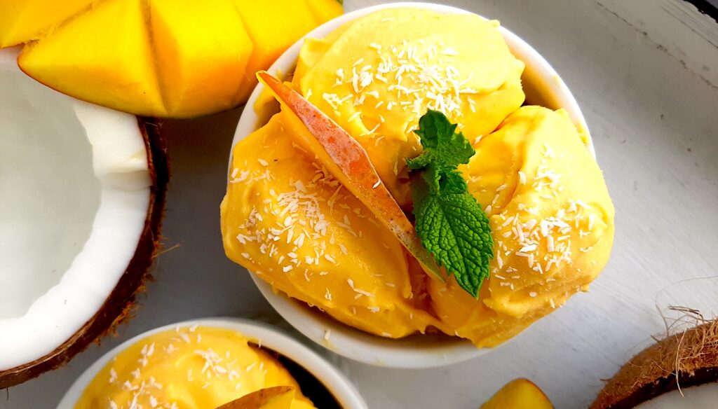 Homemade mango coconut soft serve ice cream in a white ramekin topped with shredded coconut and mint sprigs. Surrounded by halved coconut, sliced mangos and a second cup of ice cream. On a white background overhead shot. Sugar with Spice Blog.