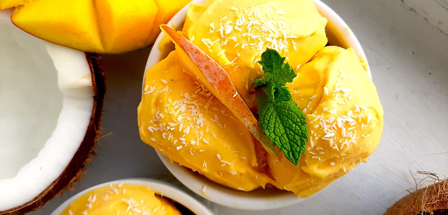 Homemade mango coconut soft serve ice cream in a white ramekin topped with shredded coconut and mint sprigs. Surrounded by halved coconut, sliced mangos and a second cup of ice cream. On a white background overhead shot. Sugar with Spice Blog.