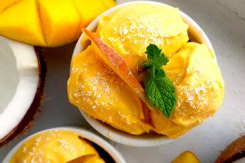 Homemade mango coconut soft serve ice cream in a white ramekin topped with shredded coconut and mint sprigs. Surrounded by halved coconut, sliced mangos and a second cup of ice cream. On a white background overhead shot. Sugar with Spice Blog.