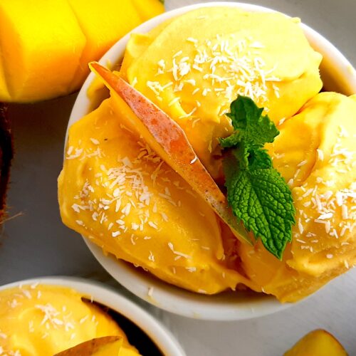 Homemade mango coconut soft serve ice cream in a white ramekin topped with shredded coconut and mint sprigs. Surrounded by halved coconut, sliced mangos and a second cup of ice cream. On a white background overhead shot. Sugar with Spice Blog.