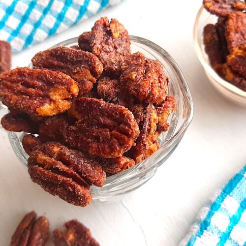 Maple candied pecans in a small glass pitcher with more maple candied pecans in a pyrex bowl behind. Blue checked dish towels nearby. A few maple candied pecans scattered on the white background. Sugar with Spice Blog.