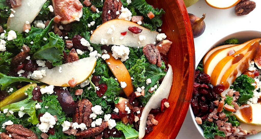 Winter Pear Salad in a tossed in a wooden bowl, ingredients separated including: sliced d'anjou pears, sliced bosc pears, gorgonzola, dried cranberries, quartered figs, sautéed pancetta, maple candied pecans, and kale. Drizzled with cranberry balsamic dressing. More winter pear salad, ingredients separated, in a white ceramic bowl to the right, half in frame. Fresh d'anjou pears, bosc pear, mission fig, maple candied pecans, and dried cranberries surround. All on a white background. Overhead shot. Sugar with Spice Blog.