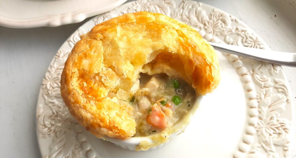 Individual chicken pot pie with puff pastry top, partially opened to reveal the pot pie, celery, chicken, carrots, and peas inside. In a white ramekin on top of a cream decorated plate with silver spoon behind. A second plate with more pies in the background. All on a white background. Sugar with Spice Blog.