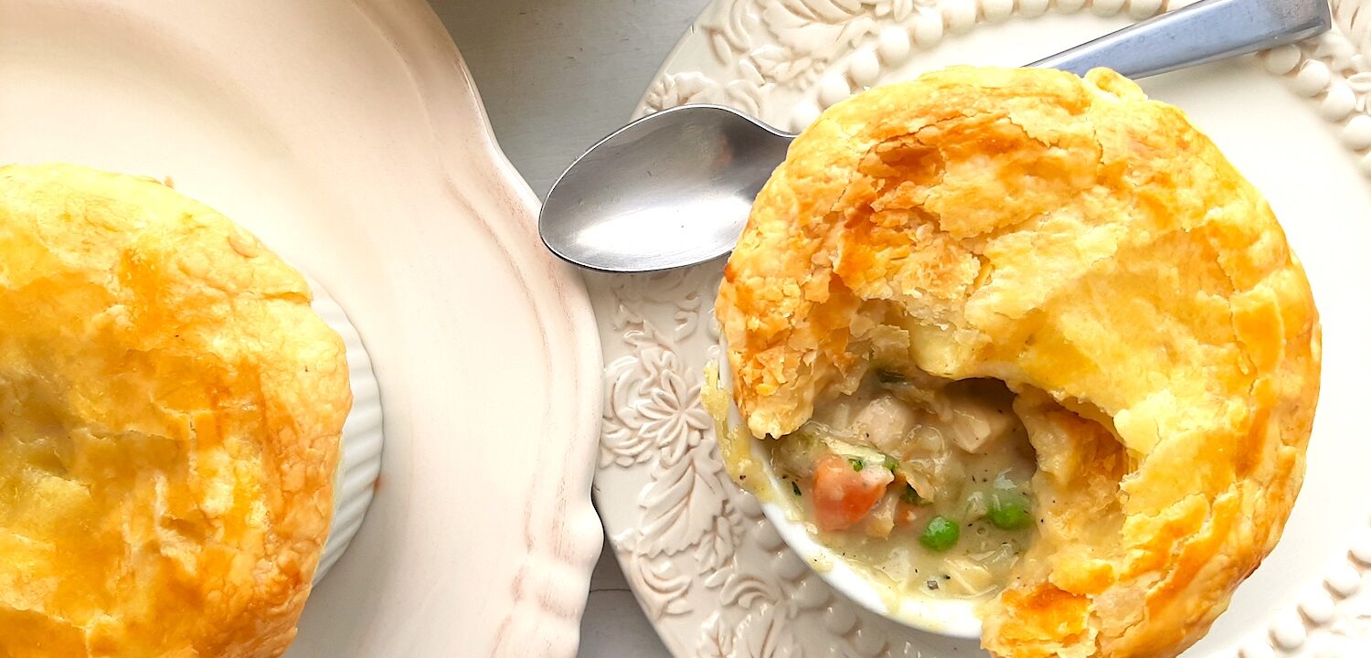 Individual chicken pot pie with puff pastry top, partially opened to reveal the pot pie, celery, chicken, carrots, and peas inside. In a white ramekin on top of a cream decorated plate with silver spoon behind. A second cream plate with more pies to the side. All on a white background. Overhead shot. Sugar with Spice Blog.