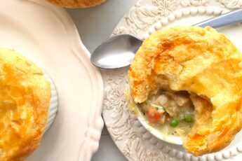 Individual chicken pot pie with puff pastry top, partially opened to reveal the pot pie, celery, chicken, carrots, and peas inside. In a white ramekin on top of a cream decorated plate with silver spoon behind. A second cream plate with more pies to the side. All on a white background. Overhead shot. Sugar with Spice Blog.