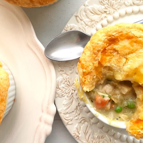 Individual chicken pot pie with puff pastry top, partially opened to reveal the pot pie, celery, chicken, carrots, and peas inside. In a white ramekin on top of a cream decorated plate with silver spoon behind. A second cream plate with more pies to the side. All on a white background. Overhead shot. Sugar with Spice Blog.