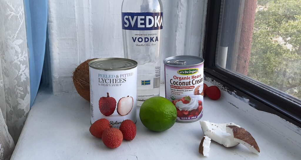 Coconut Lychee Martini Ingredients. Vodka, canned lychees in syrup, unsweetened coconut cream, fresh lychees, fresh coconut, and fresh limes on a white window sill. Front Shot. Sugar with Spice Blog.