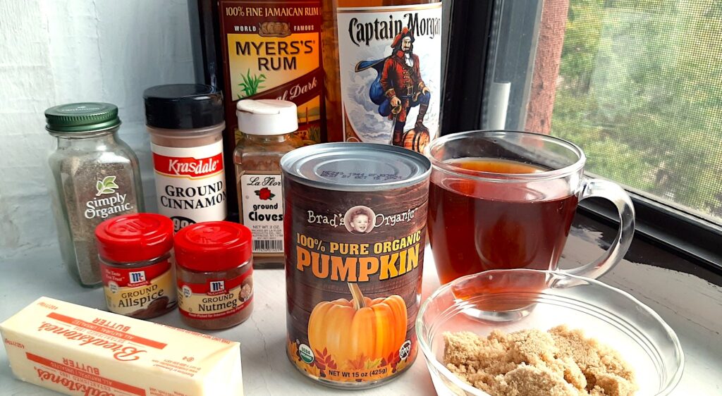 Ingredients for pumpkin spice hot buttered rum on a white window sill. Includes spiced or dark rum, canned pumpkin puree, black tea, brown sugar in a pyrex dish, 1 stick unsalted butter, ground nutmeg, cardamom, cloves, cinnamon, and allspice. Front shot. Sugar with Spice Blog.