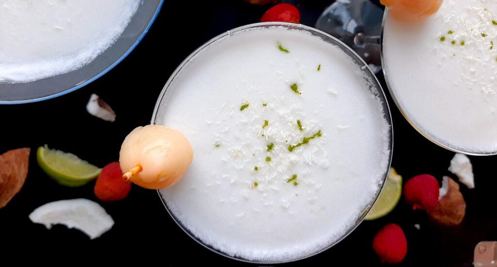 Coconut lychee martinis in three martini glasses, garnished with shaved coconut, lime zest, and a canned lychee. Fresh coconut, lime wedges, and lychees surround on a black background. Overhead shot. Sugar with Spice Blog.