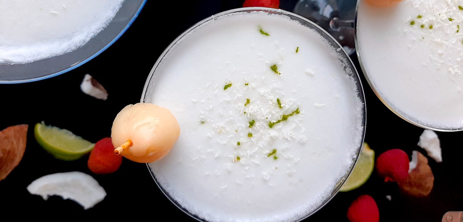 Coconut lychee martinis in three martini glasses, garnished with shaved coconut, lime zest, and a canned lychee. Fresh coconut, lime wedges, and lychees surround on a black background. Overhead shot. Sugar with Spice Blog.