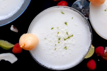 Coconut lychee martinis in three martini glasses, garnished with shaved coconut, lime zest, and a canned lychee. Fresh coconut, lime wedges, and lychees surround on a black background. Overhead shot. Sugar with Spice Blog.