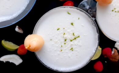 Coconut lychee martinis in three martini glasses, garnished with shaved coconut, lime zest, and a canned lychee. Fresh coconut, lime wedges, and lychees surround on a black background. Overhead shot. Sugar with Spice Blog.