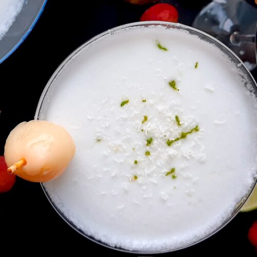 Coconut lychee martinis in three martini glasses, garnished with shaved coconut, lime zest, and a canned lychee. Fresh coconut, lime wedges, and lychees surround on a black background. Overhead shot. Sugar with Spice Blog.