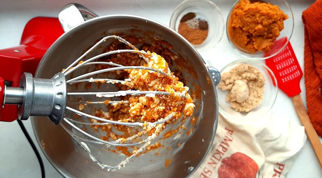 Pumpkin Spice Hot buttered rum batter being mixed in a red kitchen aid stand mixer. Pyrex dishes of brown sugar, canned pumpkin puree, and ground spices to the side with two fall-themed dish towels and a red spatula.