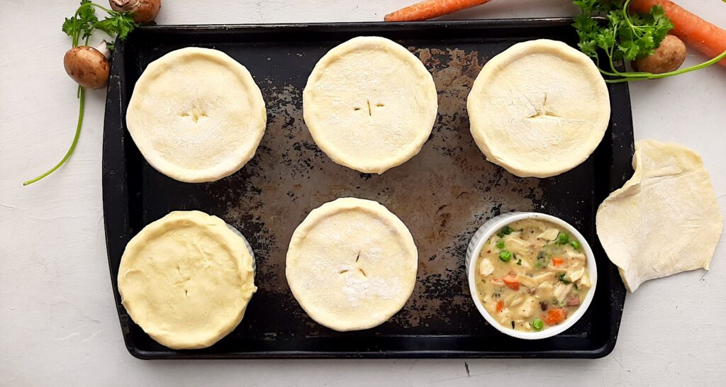 Individual chicken pot pies in white ramekins covered in puff pastry, 6 pot pies on a rusty baking sheet, one pot pie without puff pastry, pastry lying nearby, mushrooms, parsley, and carrots nearby on a white background. Overhead shot. Sugar with Spice Blog.