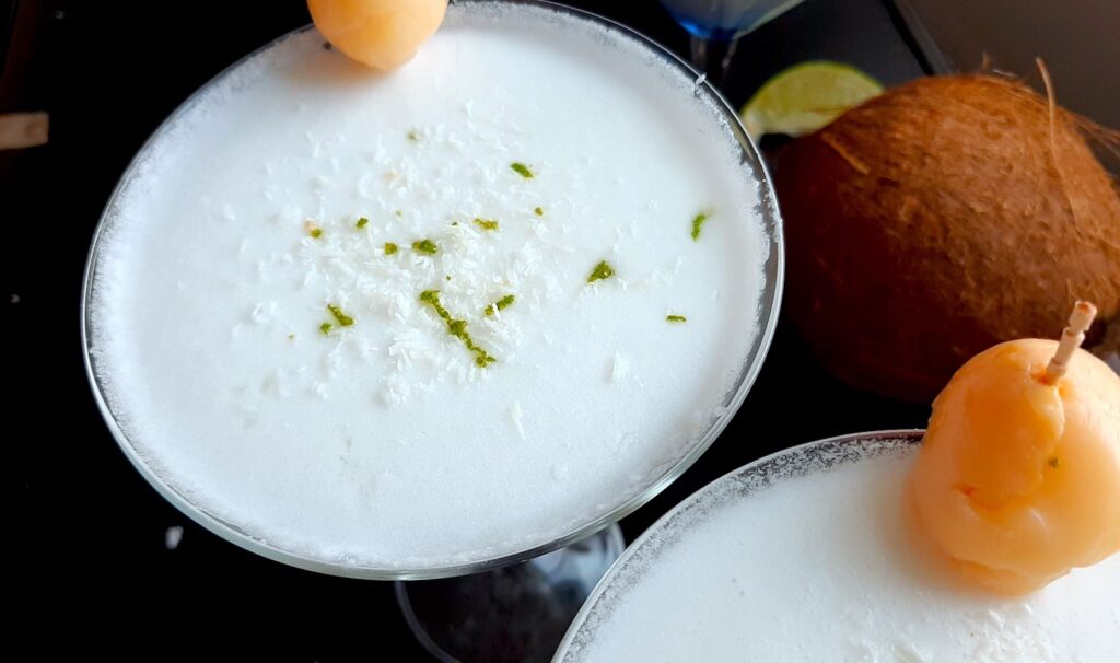 Coconut lychee martinis in two martini glasses, garnished with shaved coconut, lime zest, and a canned lychee. Fresh coconut, lime wedges, and lychees surround on a black background. Close Up shot. Sugar with Spice Blog.