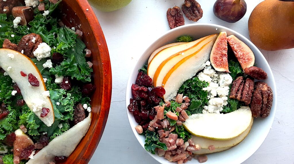 Winter Pear Salad in a white ceramic bowl, ingredients separated including: sliced d'anjou pears, sliced bosc pears, gorgonzola, dried cranberries, quartered figs, sautéed pancetta, maple candied pecans, and kale. More winter pear salad, tossed, in a wooden bowl to the left, half in frame. Fresh d'anjou pears, bosc pear, mission fig, maple candied pecans, and dried cranberries surround. All on a white background. Overhead shot. Sugar with Spice Blog.