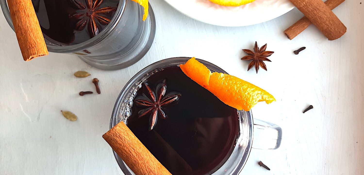 Mulled Red Wine in two clear glass mugs garnished with orange twists, star anise, and cinnamon sticks. Fresh ginger root and orange slices on a cream plate in the background. More cinnamon sticks, cardamom pods, whole cloves, and star anise scatter around on a white background. Overhead Shot. Sugar with Spice Blog.