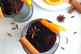 Mulled Red Wine in two clear glass mugs garnished with orange twists, star anise, and cinnamon sticks. Fresh ginger root and orange slices on a cream plate in the background. More cinnamon sticks, cardamom pods, whole cloves, and star anise scatter around on a white background. Overhead Shot. Sugar with Spice Blog.