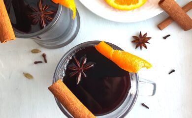 Mulled Red Wine in two clear glass mugs garnished with orange twists, star anise, and cinnamon sticks. Fresh ginger root and orange slices on a cream plate in the background. More cinnamon sticks, cardamom pods, whole cloves, and star anise scatter around on a white background. Overhead Shot. Sugar with Spice Blog.