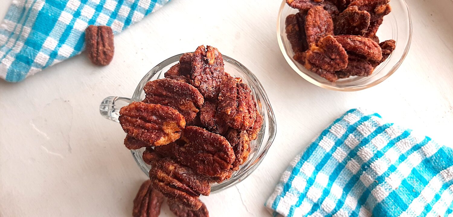 Maple candied pecans in a small glass pitcher with more maple candied pecans in a pyrex bowl behind. Blue checked dish towels nearby. A few maple candied pecans scattered on the white background. Overhead shot. Sugar with Spice Blog.