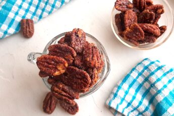 Maple candied pecans in a small glass pitcher with more maple candied pecans in a pyrex bowl behind. Blue checked dish towels nearby. A few maple candied pecans scattered on the white background. Overhead shot. Sugar with Spice Blog.