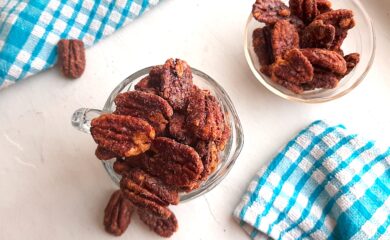 Maple candied pecans in a small glass pitcher with more maple candied pecans in a pyrex bowl behind. Blue checked dish towels nearby. A few maple candied pecans scattered on the white background. Overhead shot. Sugar with Spice Blog.