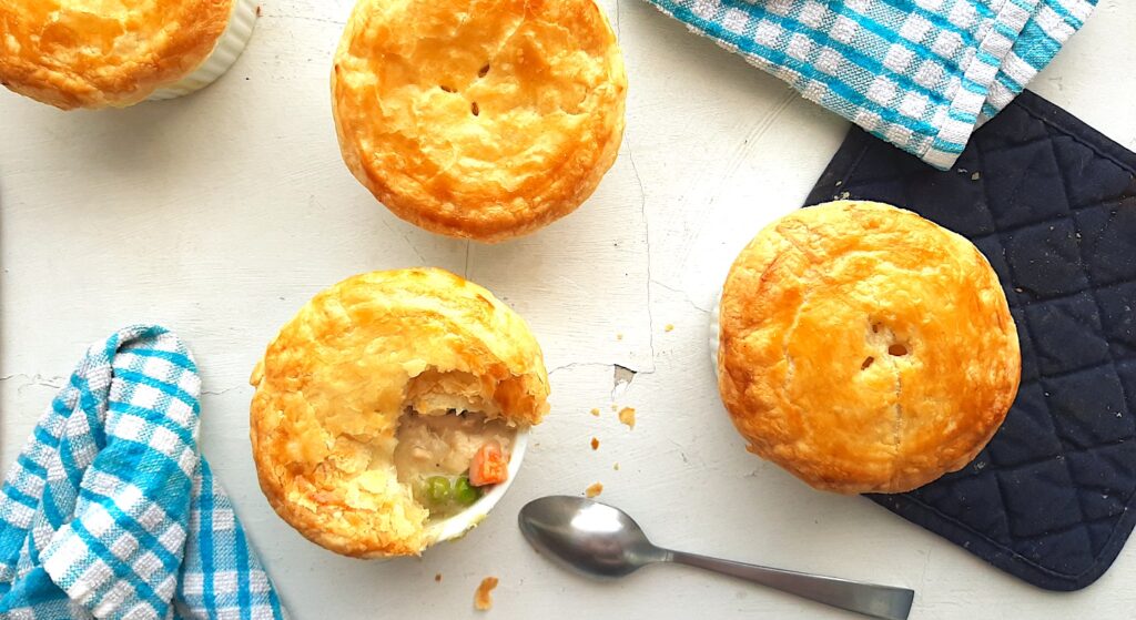 Individual chicken pot pies. Four fully cooked leftover chicken pot pies with puff pastry tops. One is opened to reveal the pot pie, peas, and carrots inside, a silver spoon lying nearby. All on a white window sill with blue checked dish towels and navy pot holders. Overhead shot. Sugar with Spice Blog.