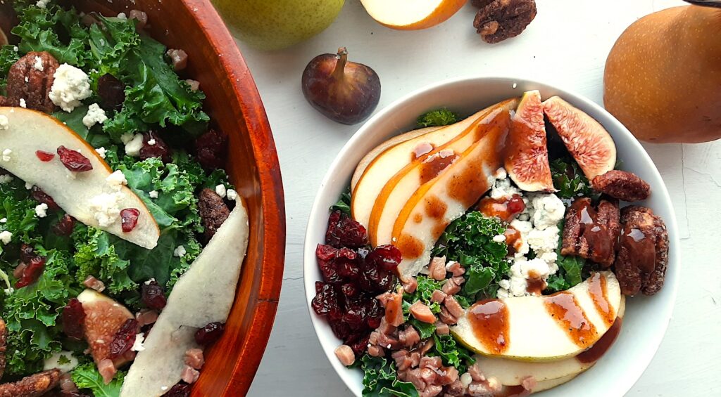 Winter Pear Salad in a white ceramic bowl bowl, ingredients separated including: sliced d'anjou pears, sliced bosc pears, gorgonzola, dried cranberries, quartered figs, sautéed pancetta, maple candied pecans, and kale. Drizzled with cranberry balsamic dressing. More winter pear salad, tossed, in a wooden bowl to the left, half in frame. Fresh d'anjou pears, bosc pear, mission fig, maple candied pecans, and dried cranberries surround. All on a white background. Overhead shot. Sugar with Spice Blog.