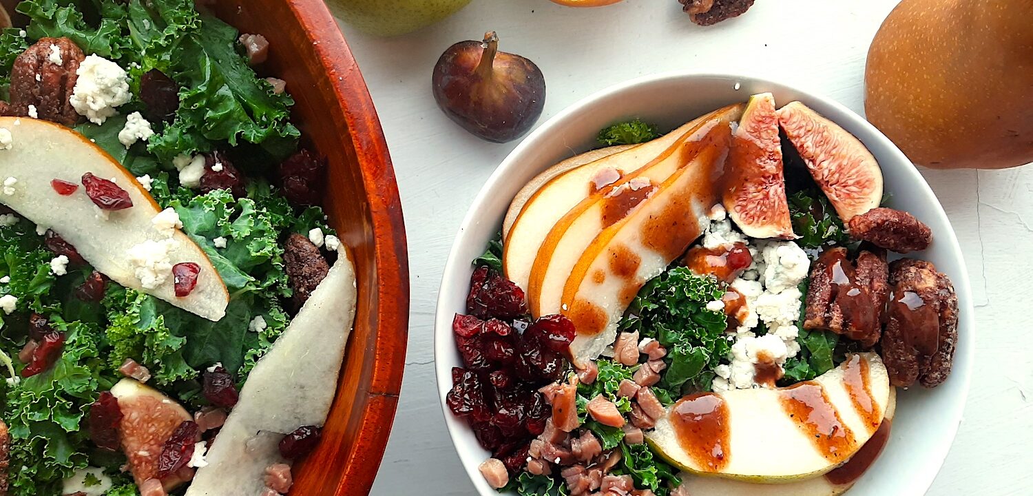 Winter Pear Salad in a white ceramic bowl bowl, ingredients separated including: sliced d'anjou pears, sliced bosc pears, gorgonzola, dried cranberries, quartered figs, sautéed pancetta, maple candied pecans, and kale. Drizzled with cranberry balsamic dressing. More winter pear salad, tossed, in a wooden bowl to the left, half in frame. Fresh d'anjou pears, bosc pear, mission fig, maple candied pecans, and dried cranberries surround. All on a white background. Overhead shot. Sugar with Spice Blog.