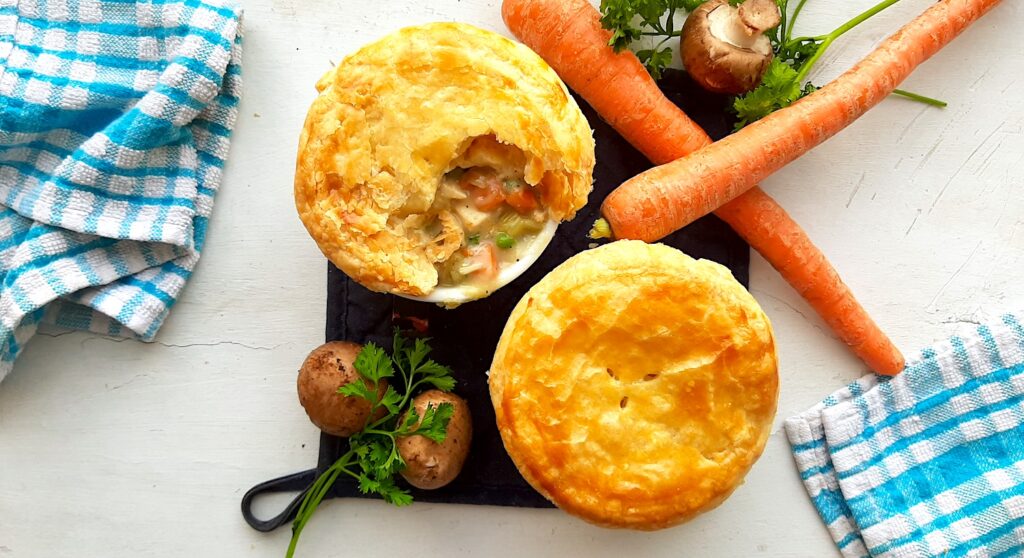Individual chicken pot pies, two fully cooked leftover chicken pot pies with puff pastry tops, one is open to reveal the pot pie, peas, and carrots inside. Both on a navy potholder with blue checked dish towels and fresh carrots, mushrooms, and parsley with blue checked dish towels nearby. All on a white background. Overhead shot. Sugar with Spice Blog.