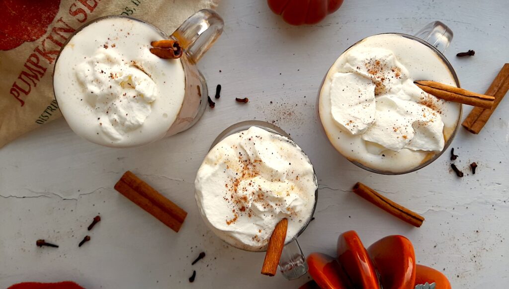 Three glasses of pumpkin spice hot buttered rum topped with whipped cream, cinnamon sticks, and ground nutmeg on a white windowsill. Window sill decorated with ceramic pumpkins, whole cloves, whole cinnamon sticks, ground nutmeg, and orange dish towels. Overhead shot. Sugar with Spice blog.