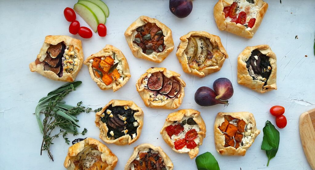 Assorted mini savory galettes surrounded by fresh herbs, vegetables, and fruits on a white background. Overhead shot. Sugar with Spice Blog.
