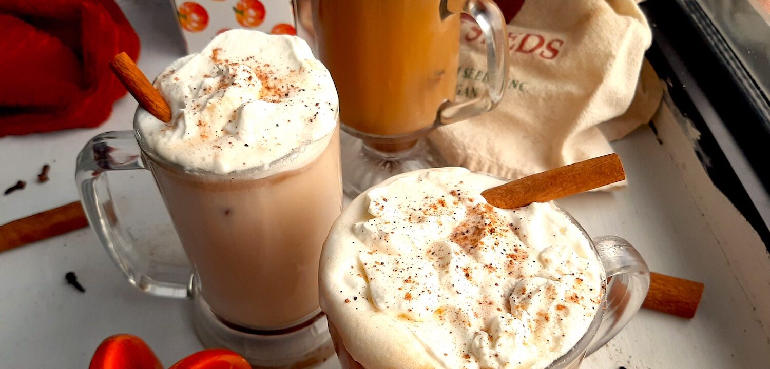 Three glasses of pumpkin spice hot buttered rum topped with whipped cream, cinnamon sticks, and ground nutmeg on a white windowsill. Window sill decorated with ceramic pumpkins, whole cloves, whole cinnamon sticks, ground nutmeg, and orange dish towels. Front Shot. Sugar with Spice blog.