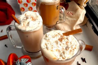 Three glasses of pumpkin spice hot buttered rum topped with whipped cream, cinnamon sticks, and ground nutmeg on a white windowsill. Window sill decorated with ceramic pumpkins, whole cloves, whole cinnamon sticks, ground nutmeg, and orange dish towels. Front Shot. Sugar with Spice blog.