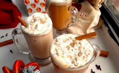 Three glasses of pumpkin spice hot buttered rum topped with whipped cream, cinnamon sticks, and ground nutmeg on a white windowsill. Window sill decorated with ceramic pumpkins, whole cloves, whole cinnamon sticks, ground nutmeg, and orange dish towels. Front Shot. Sugar with Spice blog.