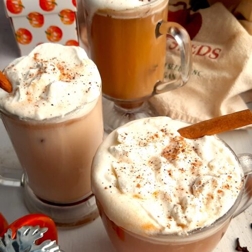 Three glasses of pumpkin spice hot buttered rum topped with whipped cream, cinnamon sticks, and ground nutmeg on a white windowsill. Window sill decorated with ceramic pumpkins, whole cloves, whole cinnamon sticks, ground nutmeg, and orange dish towels. Front Shot. Sugar with Spice blog.