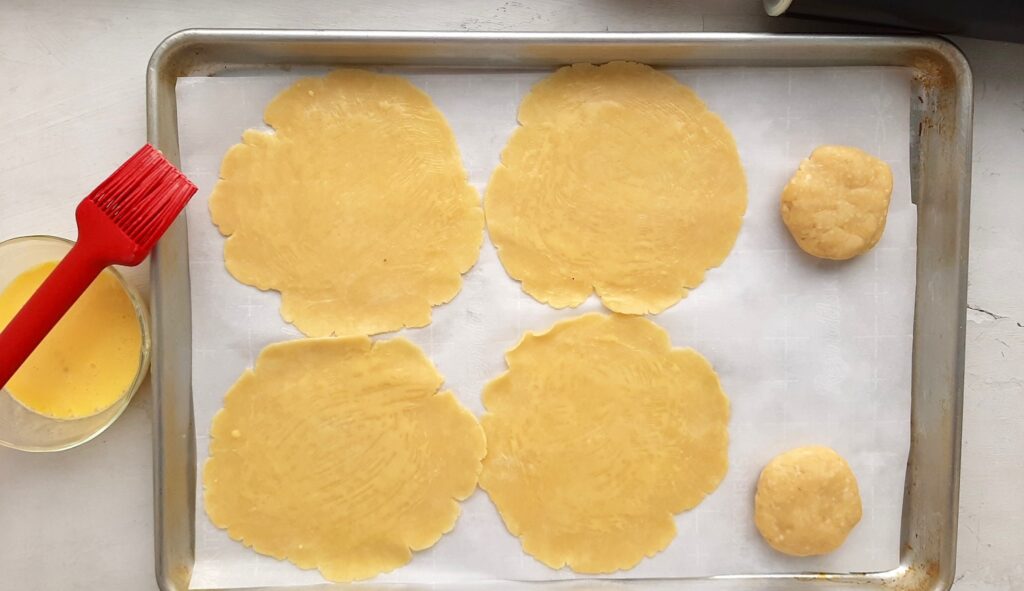 Six small raw pie crusts, four rolled out, 2 still in dough balls, on a parchment paper-lined baking tray. Overhead shot. Egg wash in a pyrex dish with red pastry brush to one side. Sugar with Spice Blog.