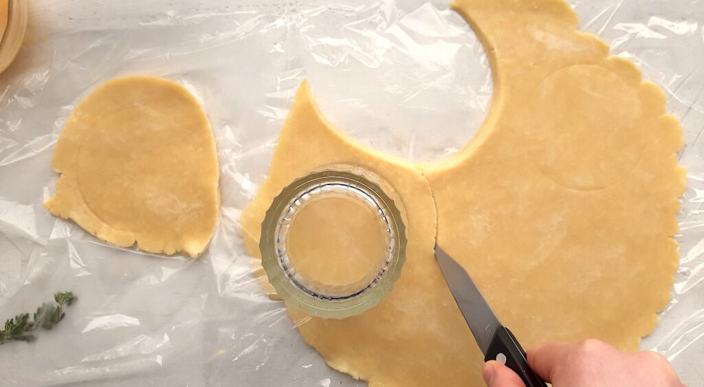 Full rolled all-butter pie crust with six small circles being measured and cut out for assorted mini savory galettes. Woman's hand uses knife and tumbler to measure and cut. Overhead shot. Sugar with Spice Blog.