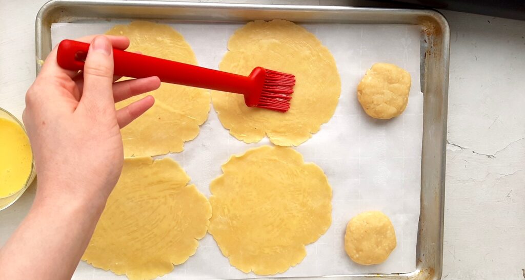 Six small raw pie crusts, four rolled out, 2 still in dough balls, on a parchment paper-lined baking tray. Woman's hand smooths egg wash with a red pastry brush over the middle crusts. Overhead shot. Sugar with Spice Blog.