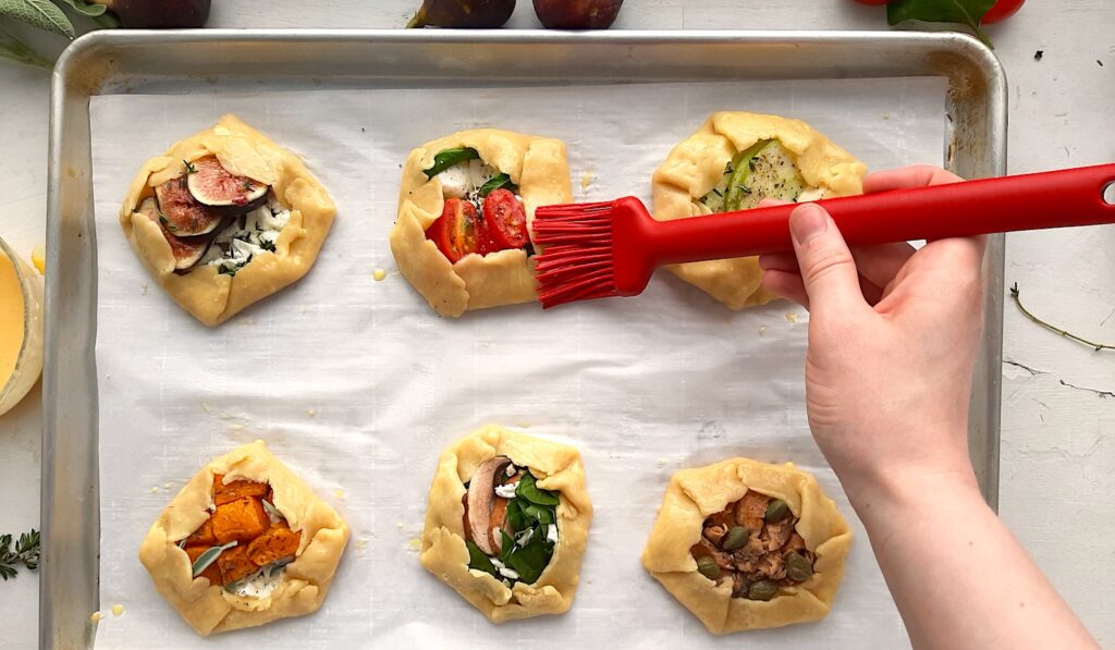 Raw assorted mini savory galettes on a parchment paper-lined baking tray. Woman's hand uses red pastry brush to smooth egg wash over the crusts. Fresh figs and sage to the side. White Background overhead shot. Sugar with Spice Blog