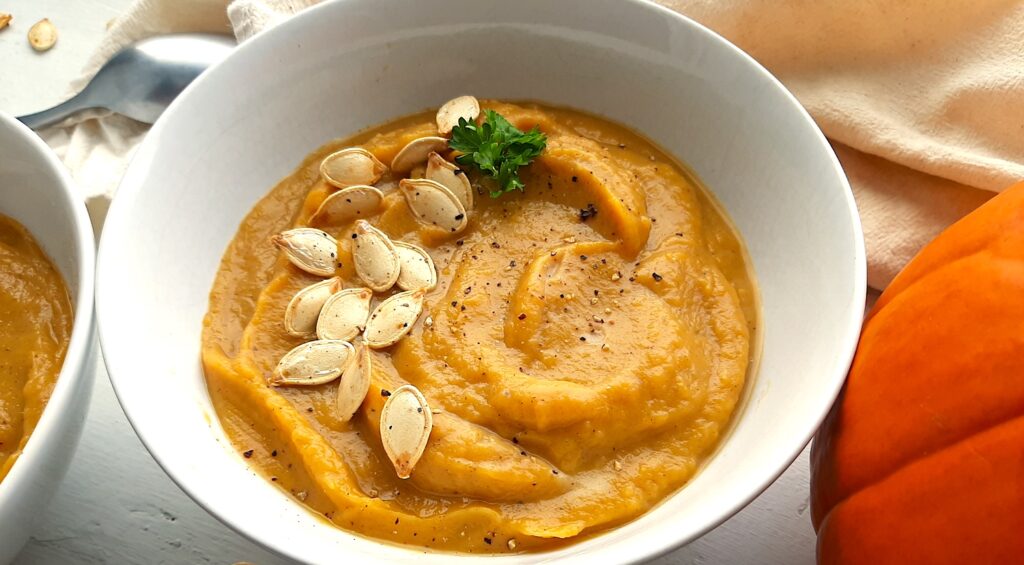Hearty roast pumpkin soup from scratch in a white bowl with pumpkin seeds, fresh parsley, and pepper to garnish. A natural fiber cloth, silver spoon, more pumpkin seeds, a second bow of soup, and a fresh pumpkin surround. Front shot. Sugar with Spice Blog.