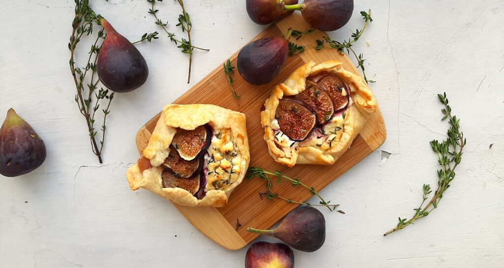 Two Mini Savory Galettes of the fig, goat cheese, thyme, and honey variety, on a wooden cutting board. Surrounded by fresh thyme and figs on a white background. Overhead shot. Sugar with Spice Blog.