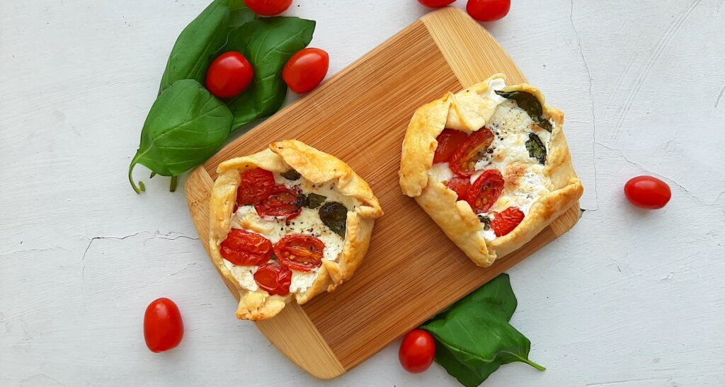 Two Mini Savory Galettes of the caprese, cherry tomato, basil, mozzarella variety, on a wooden cutting board. Surrounded by fresh basil and tomatoes on a white background. Overhead shot. Sugar with Spice Blog.