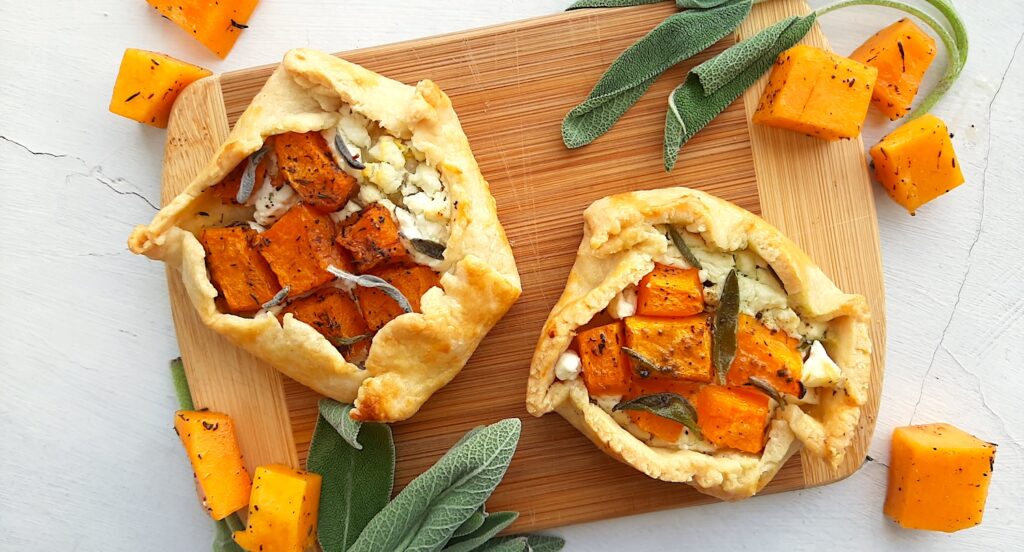 Two Mini Savory Galettes of the butternut squash, goat cheese, and sage variety, on a wooden cutting board. Surrounded by fresh sage and butternut squash cubes on a white background. Overhead shot. Sugar with Spice Blog.