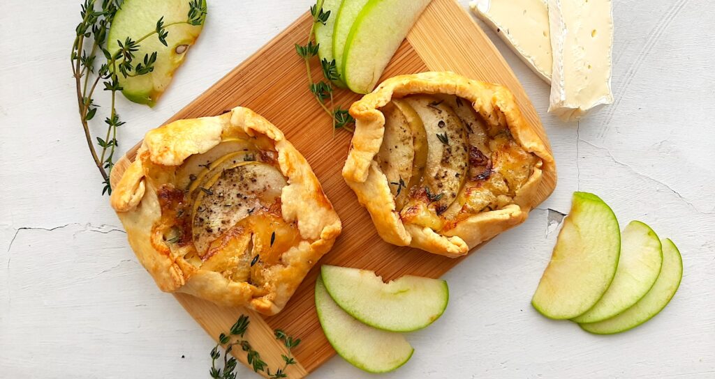 Two Mini Savory Galettes of the apple, brie, and herb variety, on a wooden cutting board. Surrounded by fresh thyme, sliced green apples and sliced brie on a white background. Overhead shot. Sugar with Spice Blog.