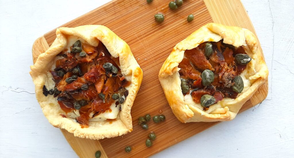 Two Mini Savory Galettes of the smoked salmon, dill, cream cheese, and capers variety, on a wooden cutting board. Surrounded by capers on a white background. Overhead shot. Sugar with Spice Blog.