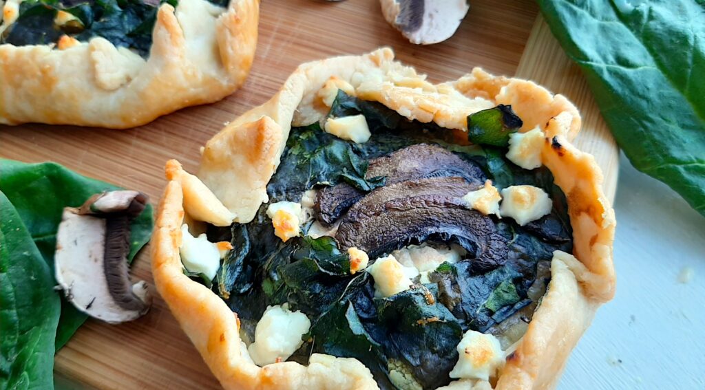 Two Mini Savory Galettes of the spinach, mushroom, and feta variety, on a wooden cutting board. Surrounded by fresh spinach and mushrooms on a white background. Front overhead shot. Sugar with Spice Blog.