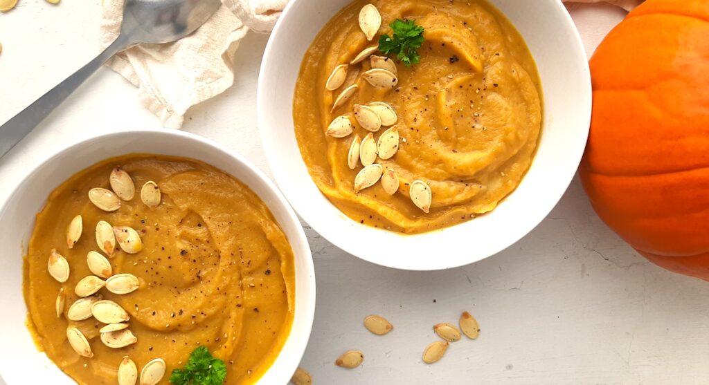 Two bowls of hearty roast pumpkin soup from scratch in white bowls, garnished with pumpkin seeds, fresh parsley sprigs, and fresh cracked pepper. A whole sugar pie pumpkin, beige cloth, silver spoon, and more pumpkin seeds are scattered around the white background. Sugar with Spice Blog.